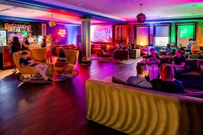 A lively lounge with colorful neon lighting, people seated on sofas and chairs around the bar area and a large screen in the back.