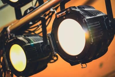 Close-up of two stage spotlights on a rig, glowing against an orange background.